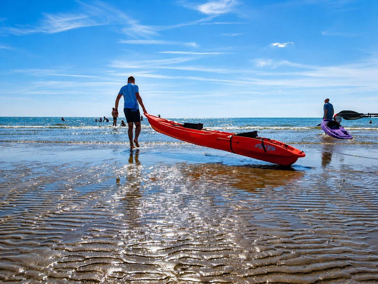 Two people taking red and purple Fatyak kayaks into the sea on a sunny beach.