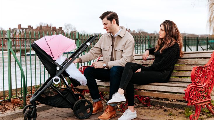 Two people sitting on a park bench beside a pink baby pushchair – For Your Little One