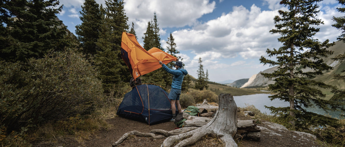 Camper setting up an orange Sierra Designs tent beside a mountain lake surrounded by trees.