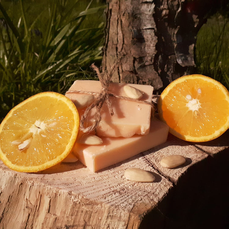 Handmade orange and almond oil soap bars by skincareBOOtique, displayed with fresh oranges on a rustic wooden log.
